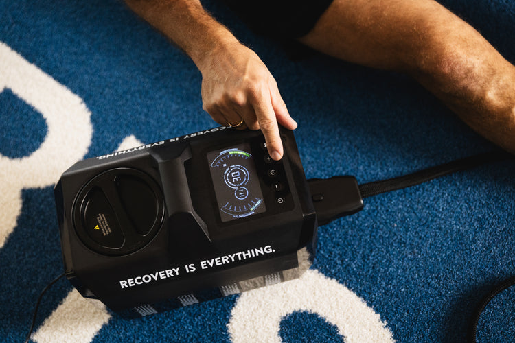 Hand bedient das Reboots Cryo One Kälte-Therapiegerät auf blauem Untergrund – modernes Recovery-System für Sportler.