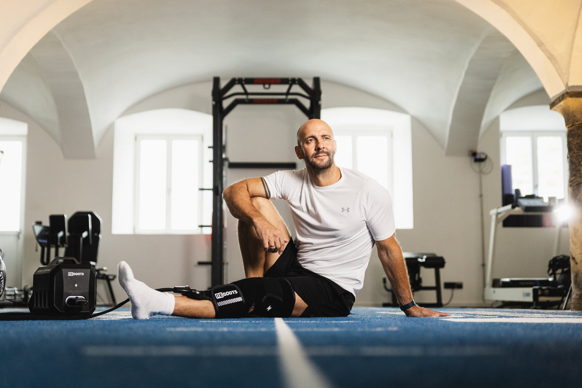 Mann sitzt im Gym am Boden und nutzt das Reboots Cryo One mit Knie-Manschette zur Kälte- und Kompressionstherapie während der Regeneration.