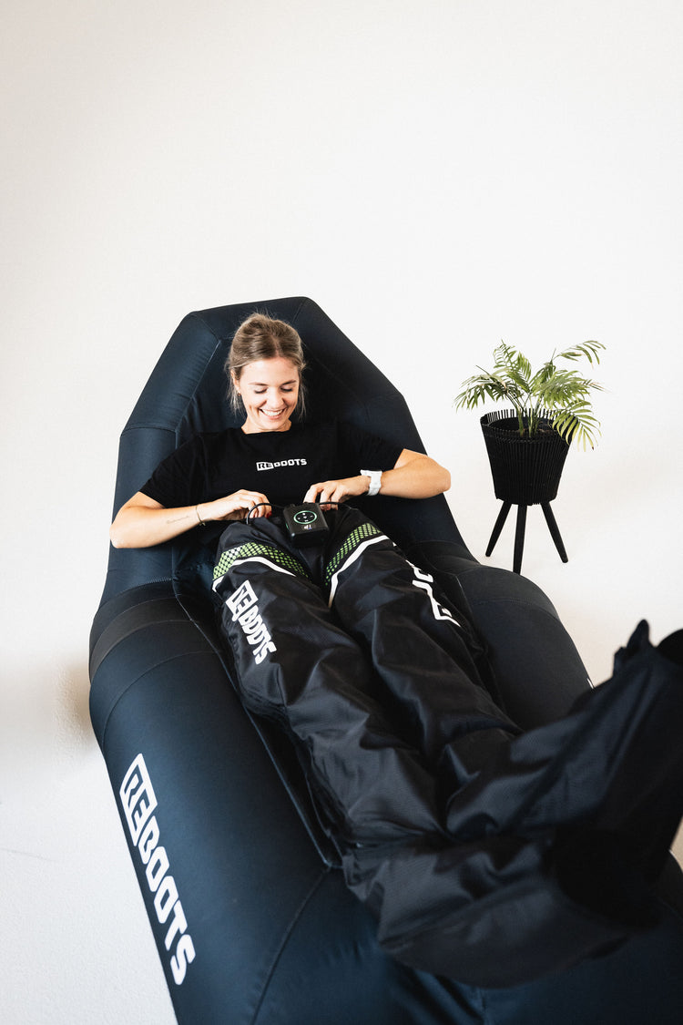 Athletin entspannt in Reboots Recovery Boots auf einer schwarzen Reboots Air Lounge und startet ihre Kompressionsmassage.
