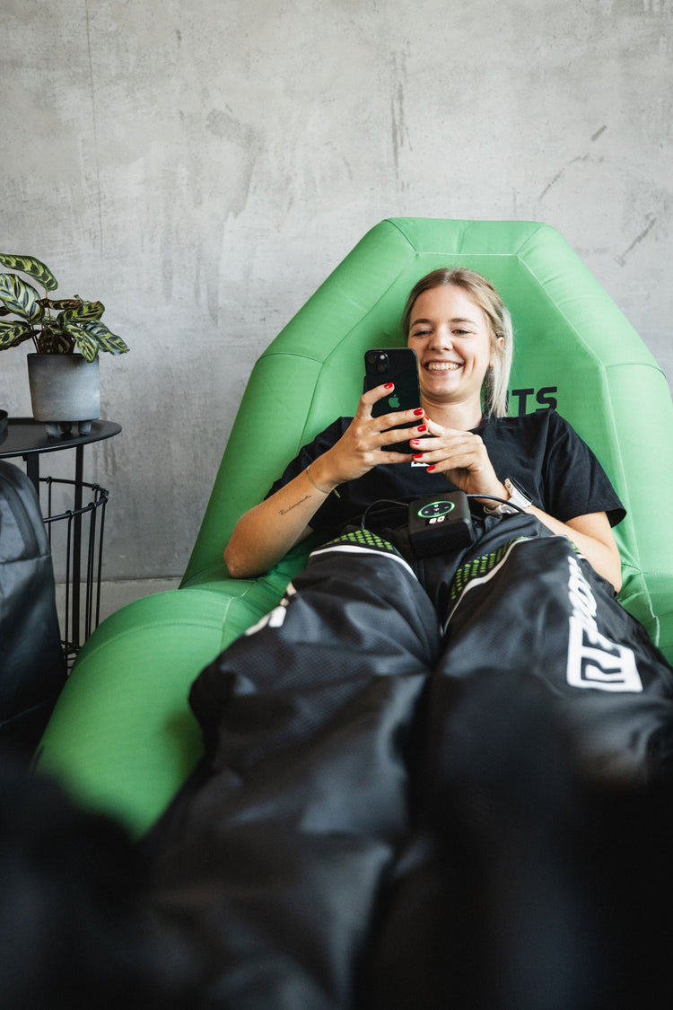 Junge Frau entspannt auf Liege und nutzt Recovery Boots zur Regeneration der Beine, während sie am Smartphone lächelt