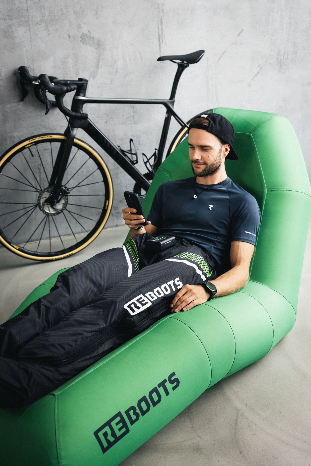 Sportler entspannt nach dem Training mit Reboots Regenerationshose auf grünem Sitzsack, Fahrrad im Hintergrund.