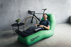 Radsportler entspannt auf grünem Reboots Recovery Chair und nutzt Reboots Kompressionsboots zur Beinregeneration, während sein Rennrad im Hintergrund steht