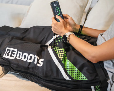 Sportlerin steuert Reboots Kompressionsboots per App und nutzt mobile Kompressionsmassage zur Regeneration bequem auf dem Sofa.